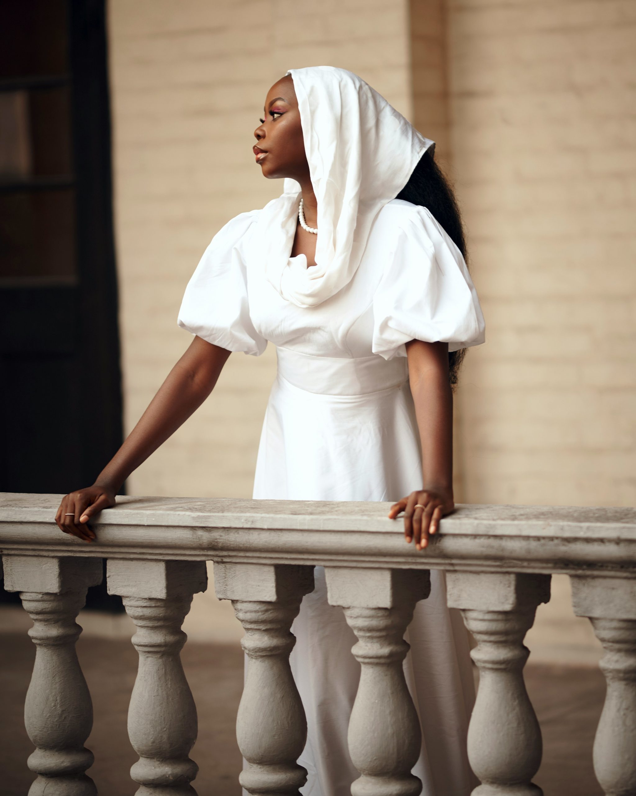 Woman in a white gown standing at a stone balustrade, poised in quiet reflection.